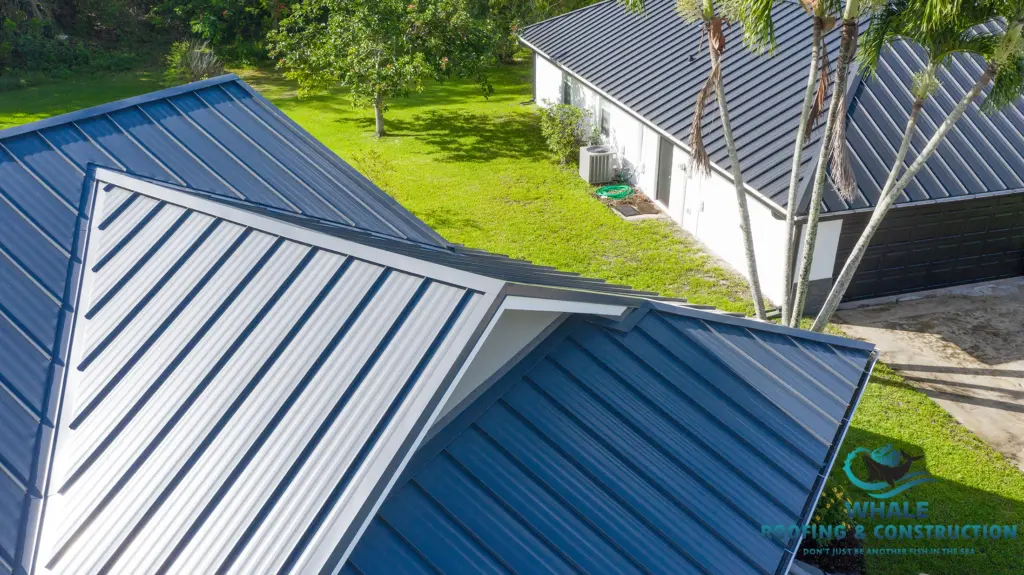 Aerial view of two buildings with dark blue standing seam metal roofs, surrounded by green grass and trees, with a visible Whale Roofing & Construction logo in the corner.