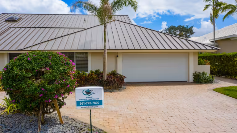 A house with a metal roof, double garage, and brick driveway displays a roofing contractor sign with a phone number in the front yard.