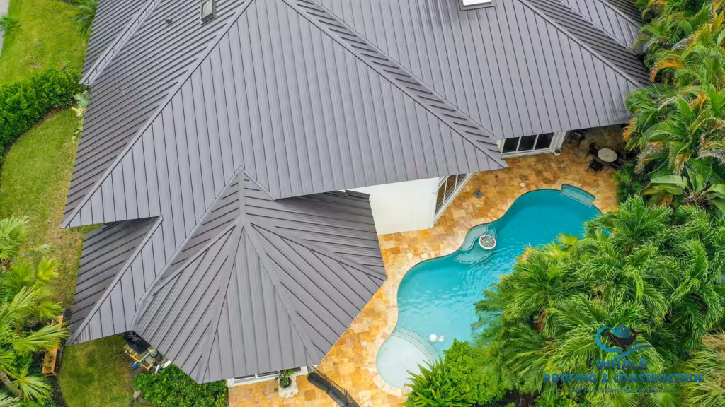 Aerial view of a house with a dark gray metal roof, stone patio, and kidney-shaped swimming pool surrounded by green trees and lawn.