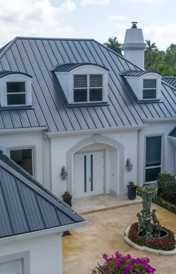 A modern house with a dark metal roof, white exterior walls, large windows, and a double-door entrance is surrounded by plants and a circular flower bed with a statue—ideal for those seeking expert roof maintenance Boca Raton services.