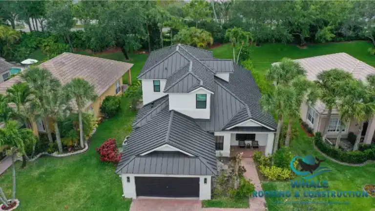 Aerial view of a modern two-story house with a dark metal roof, white exterior walls, and a brick driveway, surrounded by green lawns and palm trees.